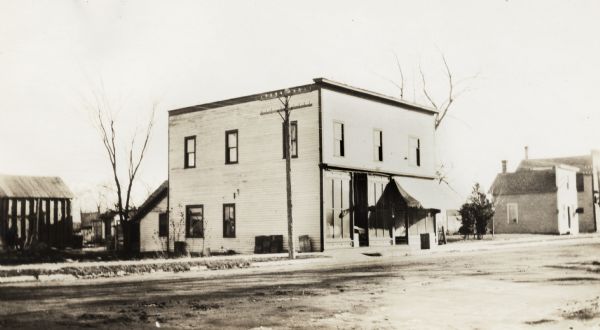 Two-story wood frame building with an awning over the front right entrance. Smaller buildings are on the right along the dirt road.