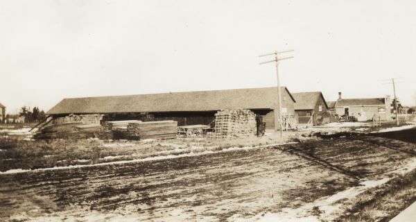 Piles of lumber are stacked on the left side of three long buildings, which are fronted along a dirt road.