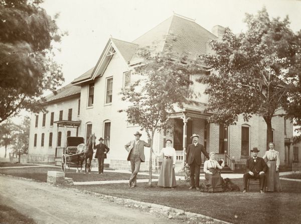 Group posing on lawn outside of the large brick building with a steep hip roof. Four men are identified (from left to right) as the turnkey, William MacKenzie, the Undersheriff, and John MacKenzie, the Sheriff.  The three women are not identified.  The man on the left is standing with a horse and buggy.  The woman sitting in a chair has a dog at her feet.