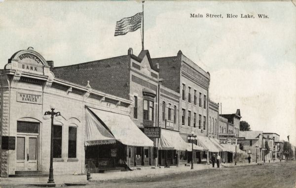 Barron Co. Bank is on the corner on the left, with storefronts lining the street going off to the right. Most of the buildings have awnings over the entrances. A flag flies from the building nest to the bank, and pedestrians are on the sidewalk. Caption reads: "Main Street, Rice Lake, Wis."