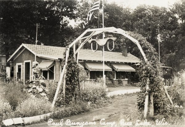 A vine-covered arched trellis with a yoke hanging from it is over the path leading into the camp. A camp building is in the background. Caption reads: "Paul Bunyans Camp, Rice Lake, Wis."