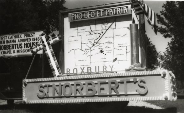 A sign above a large map of Wisconsin counties is in Latin: "Pro Deo Et Patria," meaning For God and Country, and on the left is a decorated cross, on top of a platform with a sign reading, "Roxbury / St. Norbert's." Another sign is on the back of the float.