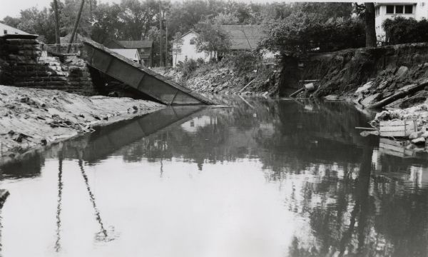 View from water of bridge collapsed into the river, with the metal supports half submerged and the brick base crumbling. Dwellings can be seen along the high banks of the shoreline.