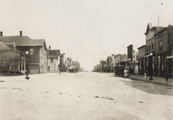 View looking down the center of road, with storefronts and dwellings on both sides. Automobiles and horse-drawn vehicles are parked at various points along the road.