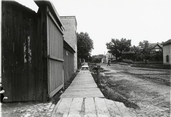 View down a wooden sidewalk, next to a dirt road. Some piles of wood are in the road near a portion of the sidewalk that is under construction.
