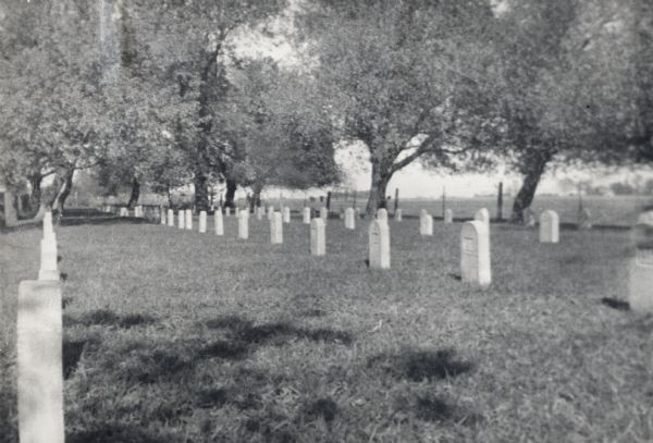 Cemetery surrounded by trees. There is a fence and field in the distance.