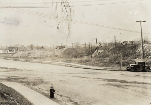 Overhead light suspended over a road, hanging from wires.  A car is parked at the corner, and billboards are in the distance. Railroad cars and railroad tracks are at the top of a small hill.