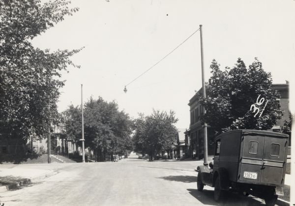 Electrical overhead street light suspended above road. A truck is parked on the right, and houses and trees line both sides of the road.