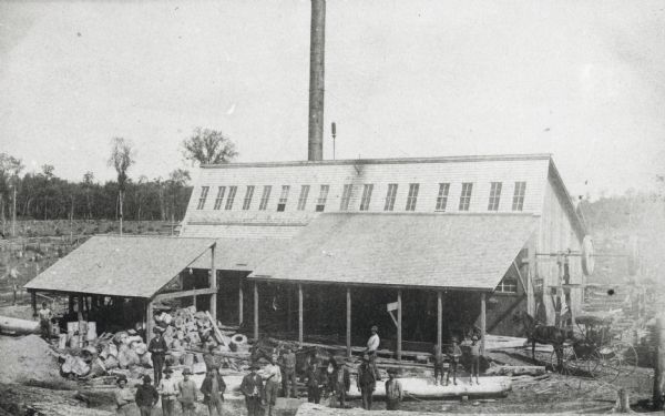 Bissell Lumber Company. Workers are posing outdoors in front of a building.  Cut trees can be seen in the background. A horse-drawn vehicle is on the right.