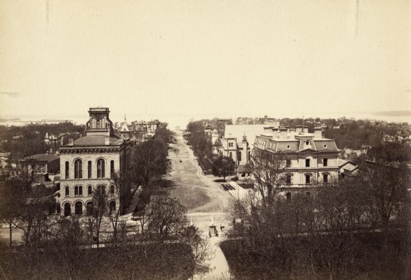 Elevated view of Wisconsin Avenue from the Wisconsin State Capitol building. Mansions and churches are along the road, and Lake Mendota and Picnic Point are in the background. City Hall is on the corner of Mifflin Street on the left.