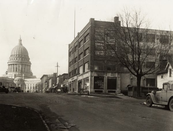 View up avenue towards the Wisconsin State Capitol building. Automobiles are parked along the street, and a building on the right has advertisements for storage.