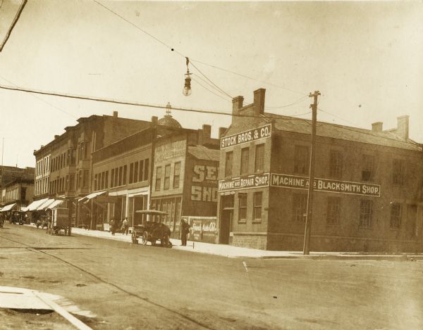 The dome is under construction and is just visible over the roof of a commercial building on East Main Street. Signs on the corner building on Main Street (to the left) at the corner of Butler Street (off to the right) say "Stock Bros. & Co.," "Machine and Repair Shop" and "Machine and Blacksmith Shop." In the street are automobiles, pedestrians, and a horse-drawn vehicle.