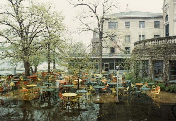 Memorial Union in the rain. Lake Mendota is visible on the left. In the foreground is the terrace area with trees and brightly colored chairs and tables. Part of the Armory (Red Gym or Old Red) can be seen in the background.