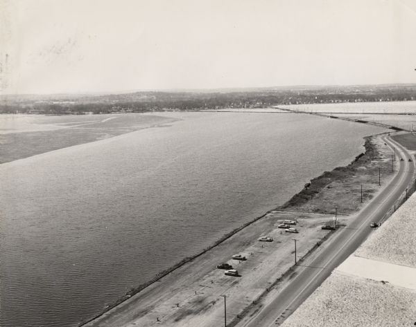Elevated view looking south over Lake Monona. Shoreline and automobiles in parking lot in lower right, next to a road. Railroad tracks cross the lake and bay in the distance. The lake is partially covered with ice.