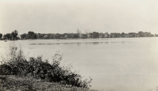 View from shoreline of tree-lined distant shore, beyond which can be seen the dome of the Wisconsin State Capitol, and on the right, St. Raphael's Catholic Church.