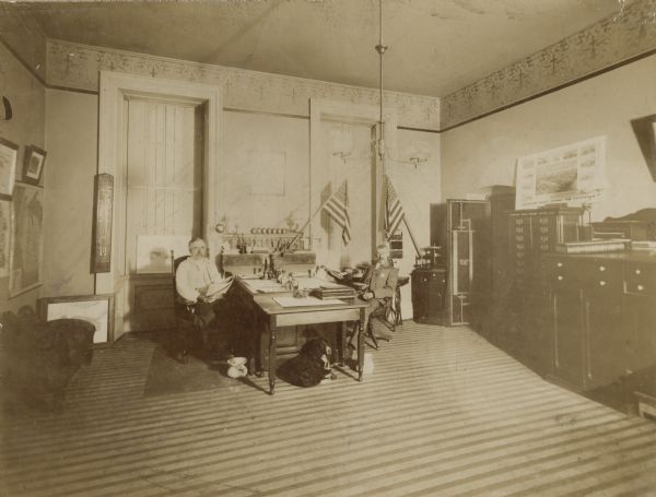 Mendota State Hospital office. Charles Carter, steward, seated at his desk.  Across from him is George B. Merrick, Asst. Steward.  Below the desk is a dog, Carlo, "Second Asst."  There is a spittoon under the desk, flags displayed along the wall, a heavy open door with lock mechanisms on the inside, and large cabinets on the right. (Mendota Mental Health Institute)