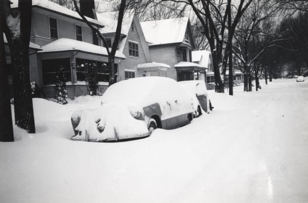 View from street of houses on the south side of Madison. The cars parked in front of the house are covered in snow.