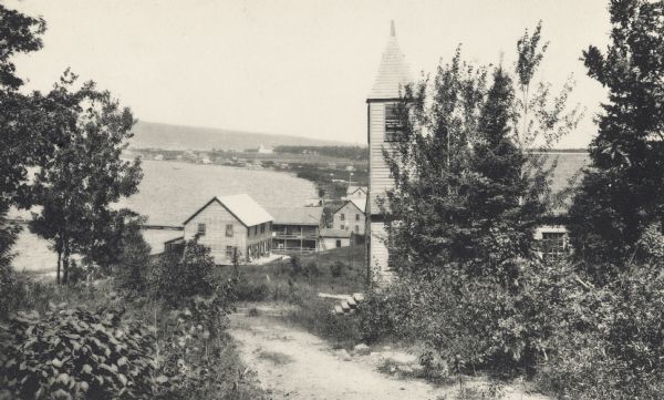 Hilltop view of the Congregational church on Madeline Island (?).  In the background are buildings, houses, and the shoreline of a lake.