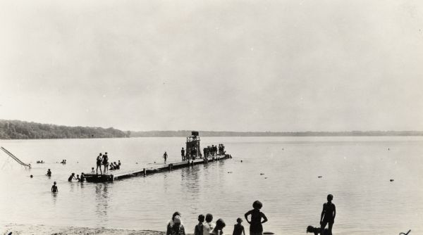 View from beach of people swimming. Some are standing in the sand, and some are on the diving pier out in the water. On the left is part of a water slide.