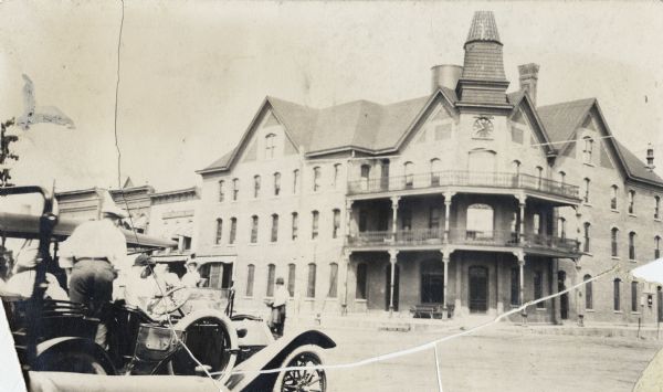 View from street of hotel with balcony above entrance on corner, and a clock tower. Motorcar with several people are on the left.
