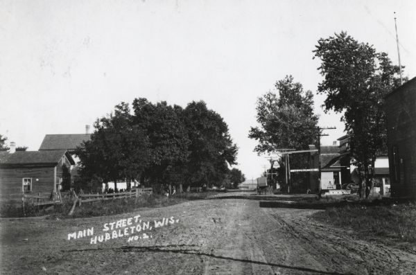 One time location of Mary Hoffard house. View down dirt road, lined with houses, shops, and trees. A horse-drawn vehicle is going down the road.