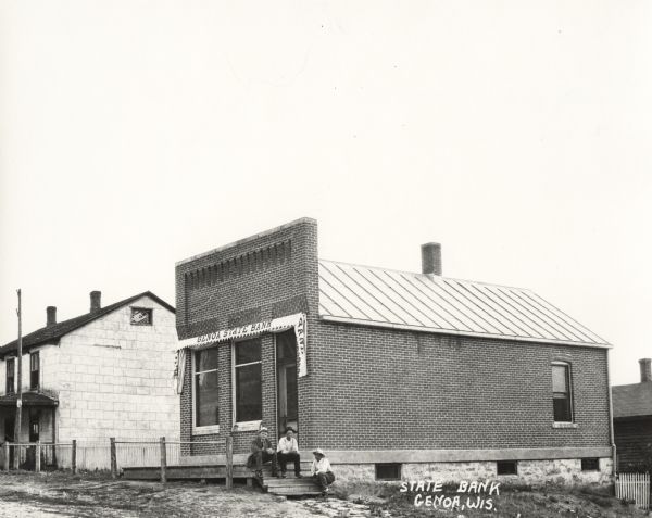 Exterior of brick building. A banner hangs over the entrance, and three men wearing hats sit on the steps near the front door. Another building is on the left.
