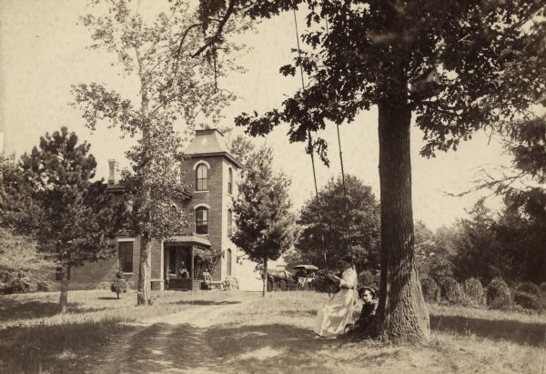 A brick house with front porch and tower sits back from a road surrounded by trees and bushes. There is a horse-drawn carriage on the drive.  Two people are in the carriage, and three are on the porch.  In the foreground, three children are on or near a swing hanging from a tree.