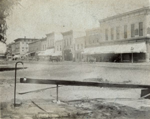 View from corner of storefronts. Horse-drawn vehicles and pedestrians are in the street. There is a water fountain with a cup in the foreground.