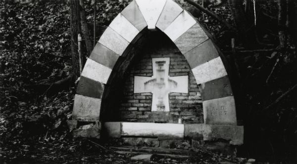 Roman Catholic grotto with a cross under a peaked arch surrounded by trees and shrubs.