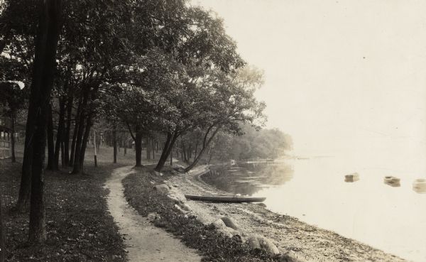 Path on tree-lined shoreline with rowboats. A section of a porch is on the left, other dwellings and piers in the distance.