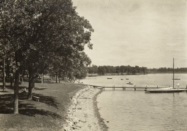 Lake Lawn. Several boats are on the water with a man standing on his sailboat next to the dock.  Trees run along the shoreline into the distance.