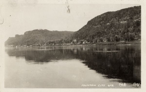 View from river of Fountain City's shoreline with bluffs in the background. Caption reads: "Fountain City, Wis."