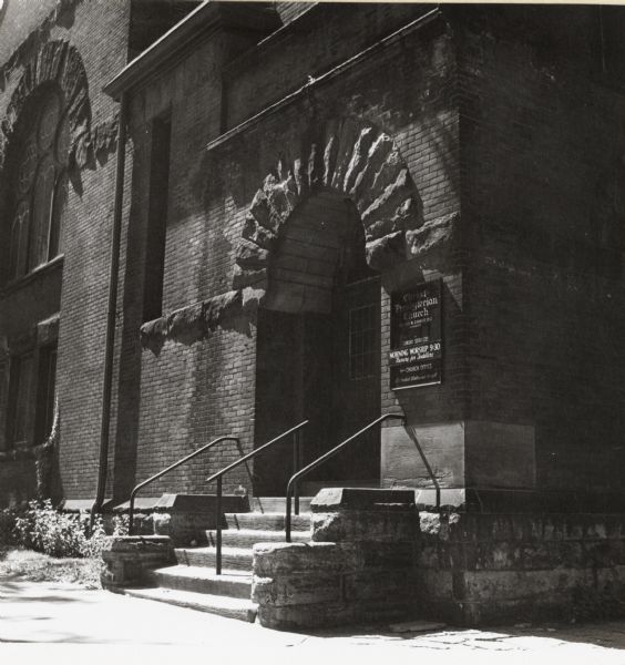 Arched entrance at corner of the building near sidewalk. Sign to the right of the door gives the times for mass.