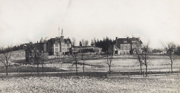 University of Wisconsin-Madison College of Agriculture, looking southeast. Fences and fields are in the foreground. There are two windmills near buildings and greenhouses on the left, the Washburn Observatory is behind them on a hill. The dome of Bascom Hall is in the background. In the center is Hiram Smith Hall, and on the right on a hill is the College of Agriculture.
