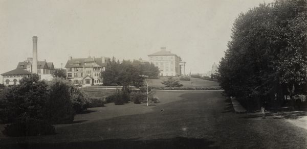 Exterior view from west of University of Wisconsin-Madison College of Agriculture. Trees and a sidewalk line the open lawn on the right.  Several buildings and a chimney are on a hill in the middle and left, including Hiram Smith Hall. On the right in the distance is Bascom Hall.