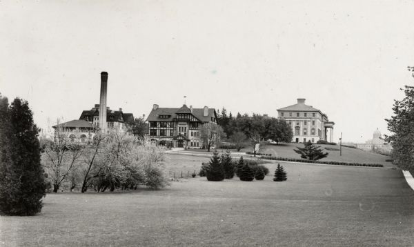 Exterior view from west of University of Wisconsin-Madison College of Agriculture. Trees and a sidewalk line the open lawn on the right. Several buildings and a chimney are on a hill in the middle and left, including Hiram Smith Hall. On the right in the distance is Bascom Hall.