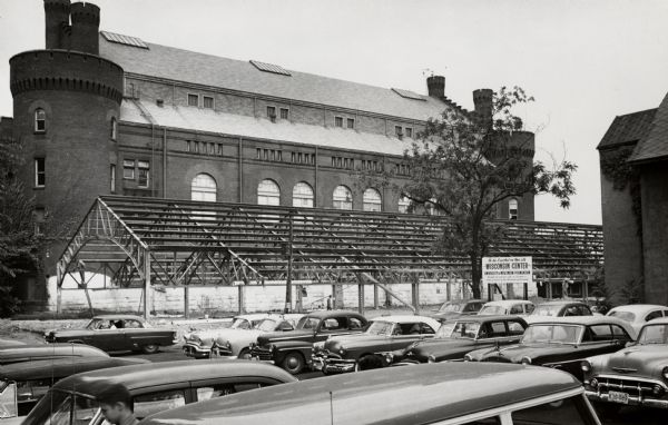 View across parking lot of Armory (Red Gym or Old Red) on the University of Wisconsin-Madison campus.  The gymnasium annex in course of demolition.  There is a sign telling of the future site for the Wisconsin Center. Men are working under the metal framework.