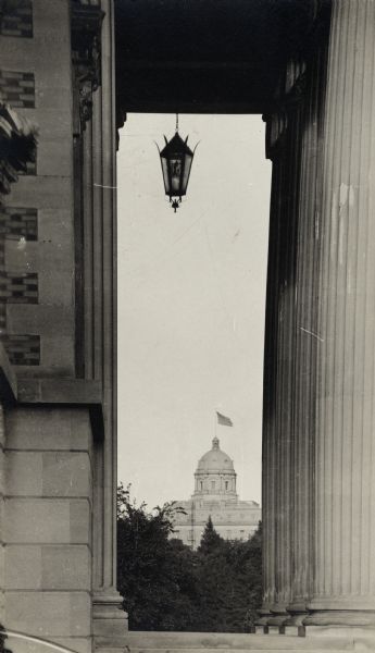 View of Bascom Hall (formerly Main Hall) through the columned entrance of the main building of the College of Agriculture. A flag is flying from the dome of Bascom Hall.
