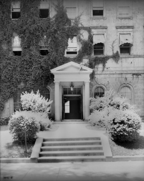 Columned side entrance, with flight of steps flanked by bushes, of Bascom Hall (formerly Main Hall) on the University of Wisconsin-Madison campus. Vines are growing on the facade of the building.