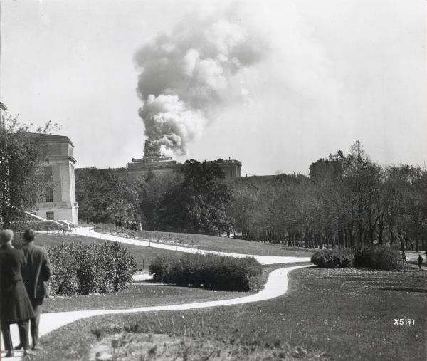 View from distance of Bascom Hall (formerly Main Hall) dome burning on the University of Wisconsin-Madison campus. Smoke is billowing out from the building, and two men on a path are watching on the left.