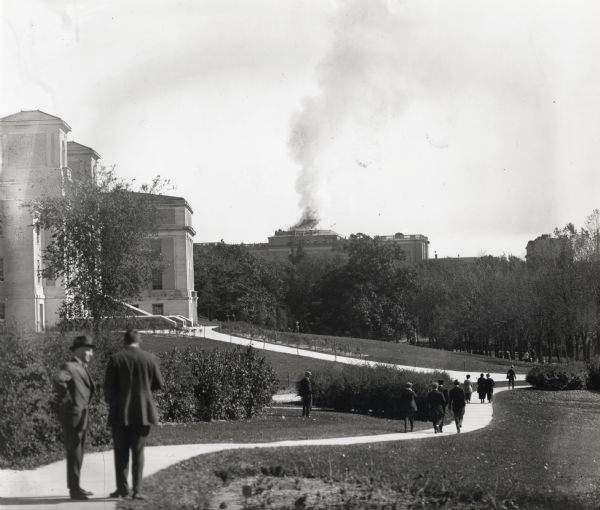 View from distance of Bascom Hall (formerly Main Hall) dome burning on the University of Wisconsin-Madison campus. Smoke is billowing out from the building, and two men on a path are watching on the left.