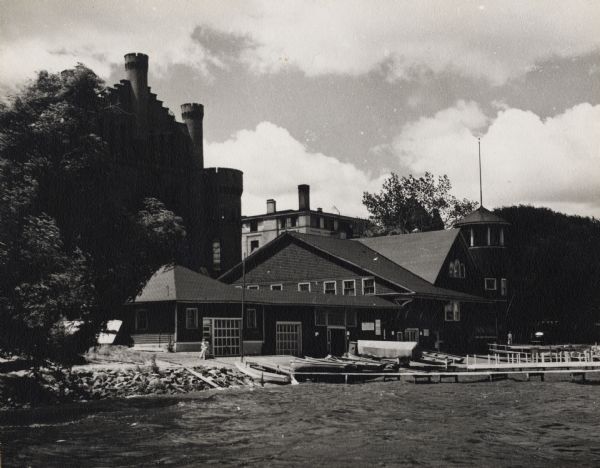 View from Lake Mendota of boathouse on the University of Wisconsin-Madison campus. The Armory (Red Gym or Old Red) is behind the boathouse. A person is sitting near boats which are stored near piers on the shoreline.