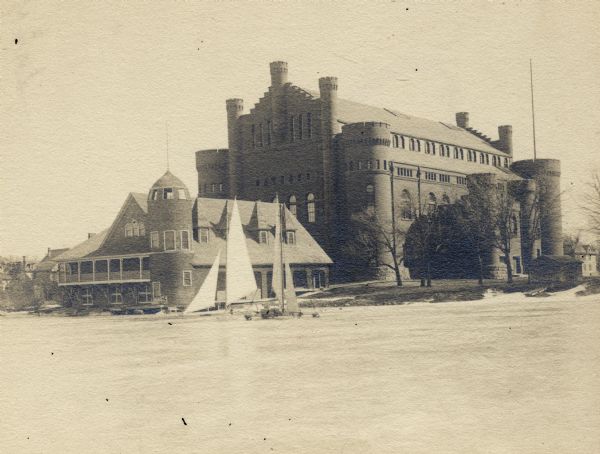 View from frozen Lake Mendota of boathouse on the University of Wisconsin-Madison campus. The Armory (Red Gym or Old Red) is behind the boathouse.  Two iceboats with sails are parked on the ice in front of the boathouse near the shoreline.
