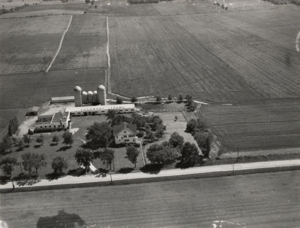 Aerial view of farm which is part of the University of Wisconsin-Madison. In the foreground is a tree-lined road. The farm has several buildings, including silos, and a long building with a sign for Charmany Farms on the roof. The farm is surrounded by fields.