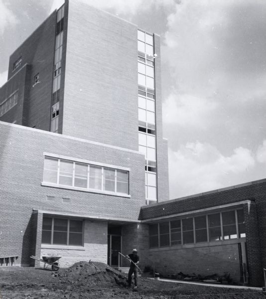 E.B. Fred Hall, the first home of the bacteriology department, on the University of Wisconsin-Madison campus. A person is shoveling dirt in front of the building, and a wheelbarrow and a ladder stand near the wall on the left.
