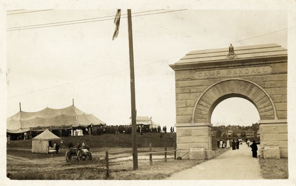 Day of the dedication of the Memorial Arch for Camp Randall on the University of Wisconsin-Madison campus. University Heights, with the Buell and Gilmore houses, is visible in the distance. A large tent is on the left with a large crowd. Other small tents are nearby.