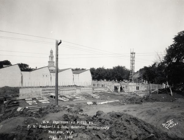 Mechanical Engineering Building under construction. A group of construction workers are on the foundation are working near a tower. There is a church steeple in the background.