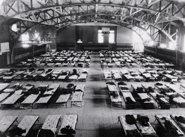 Elevated view of rows of cots in the Armory (Red Gym or Old Red) building at the University of Wisconsin-Madison. Dormitory installed in Men's Gym. A raised platform on the left has chairs and tables.