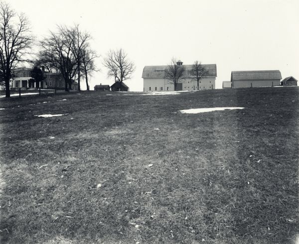View from field of farm buildings of the University of Wisconsin-Madison. Beyond a partially snow-covered field are several barns, and a large home to the left.