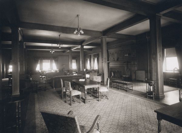 Reception Room of hall at University of Wisconsin-Madison campus. A large fireplace is along the right wall, and decorative pillars support the room. Tables, chairs, and couches are arranged around the room.